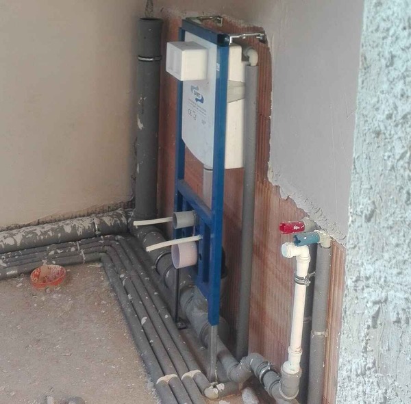 Montowana na ścianie rama toalety z odsłoniętą instalacją wodno-kanalizacyjną i rurami jest częściowo zainstalowana w niewykończonym pomieszczeniu z gołymi ścianami i widocznymi materiałami budowlanymi.