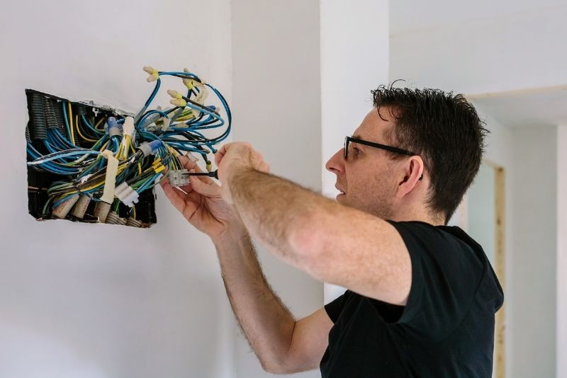 Osoba w okularach i czarnej koszuli umiejętnie pracuje nad okablowaniem elektrycznym wewnątrz ściany, postępując zgodnie z instrukcjami. Odsłonięte przewody wyświetlają różne kolory, gdy używają narzędzia do regulacji połączeń w tym wewnętrznym otoczeniu.