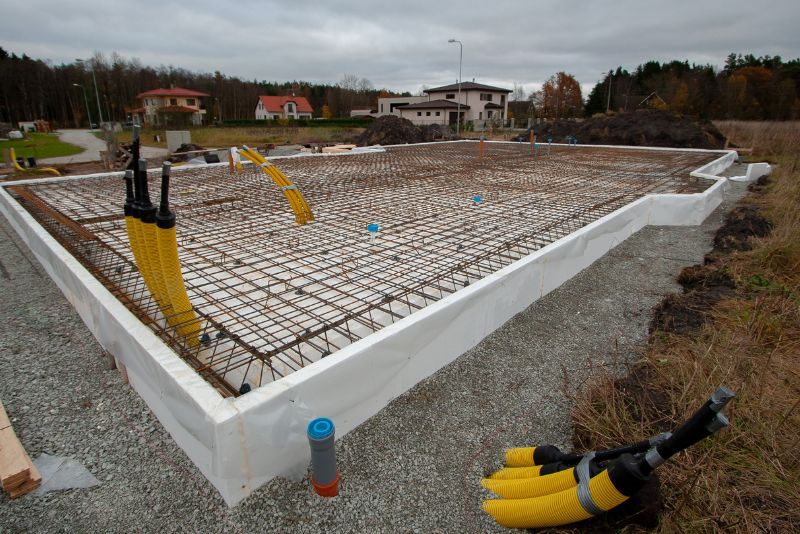 Betonowy fundament w trakcie budowy z siatką stalowych prętów zbrojeniowych, białą folią z tworzywa sztucznego, żółtymi rurami i niebieskimi odgałęzieniami przewodów. W tle domy mieszkalne i drzewa pod zachmurzonym niebem.