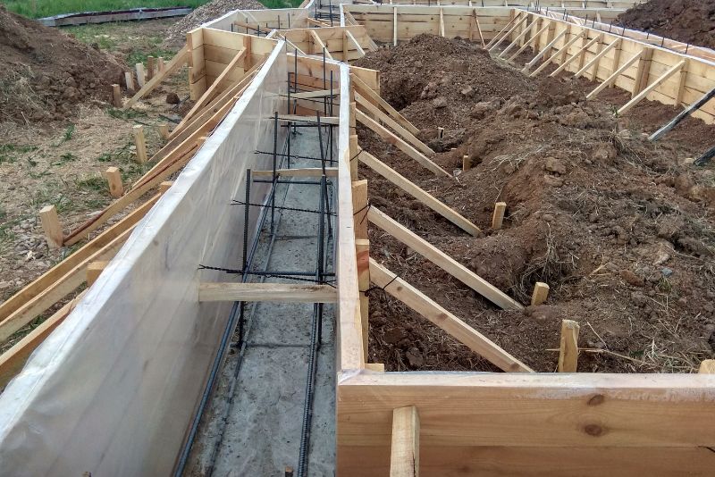 Drewniany szalunek i stalowe pręty zbrojeniowe są ustawione pod betonowy fundament, z widocznym gruntem i wspornikami.