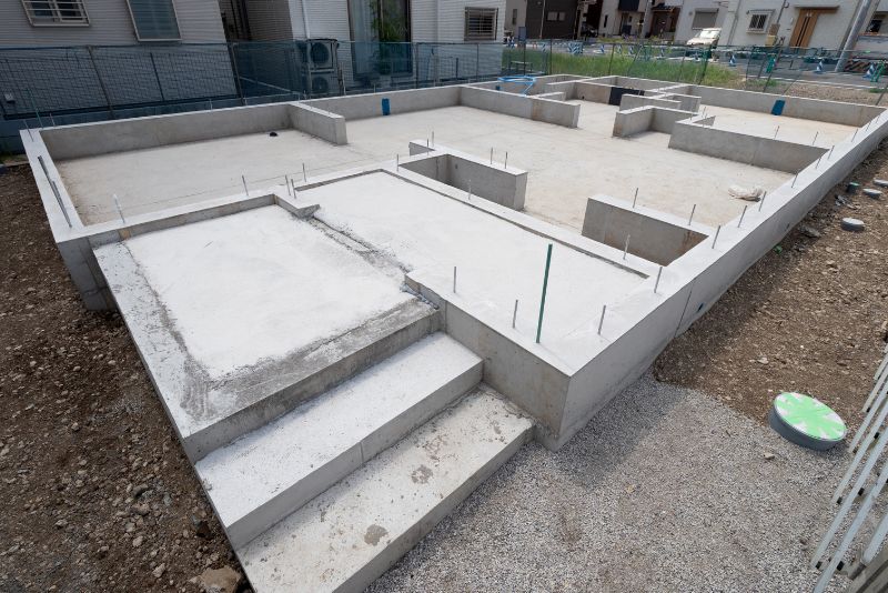 Betonowy fundament domu ze stopniami, widocznymi prętami zbrojeniowymi i prostokątnymi kształtownikami osadzony w ziemi. Otoczony ogrodzeniem i sąsiednimi budynkami mieszkalnymi w tle.