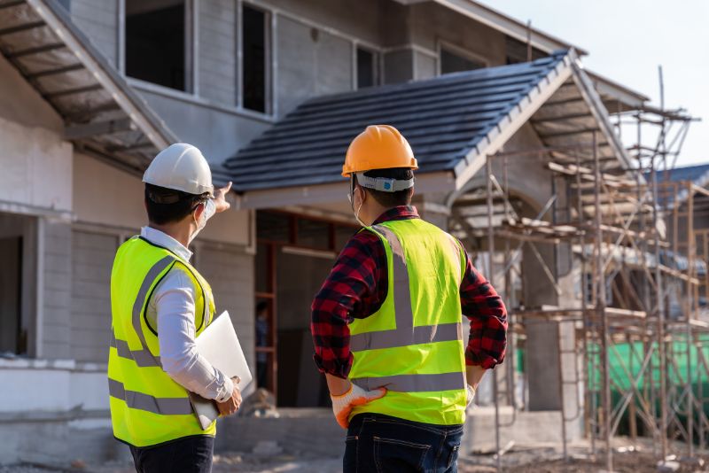 Dwóch pracowników budowlanych w kaskach i odblaskowych kamizelkach stoi naprzeciwko niedokończonego budynku. Jeden wskazuje na konstrukcję, podczas gdy drugi trzyma plan, a rusztowanie przedstawia ich ambitny projekt.