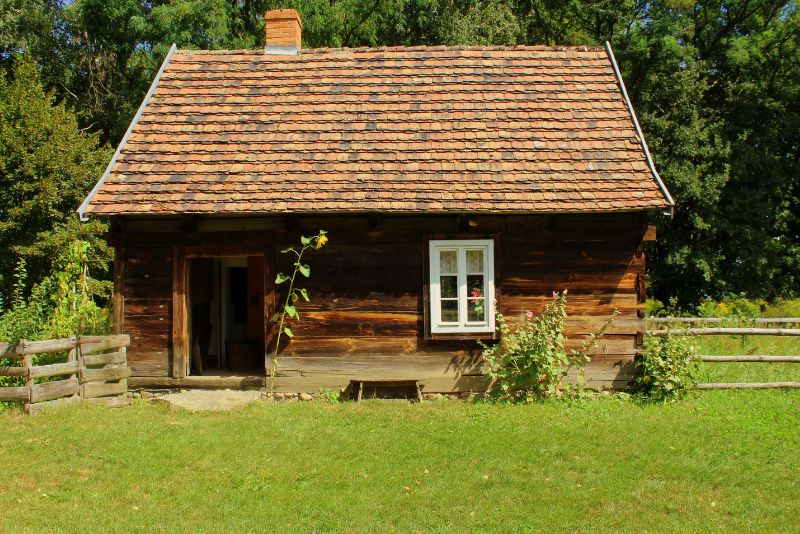 Mały drewniany domek z dachem krytym dachówką, pojedynczym oknem i otwartymi drzwiami stoi za płotem wśród zieleni w słoneczny dzień.