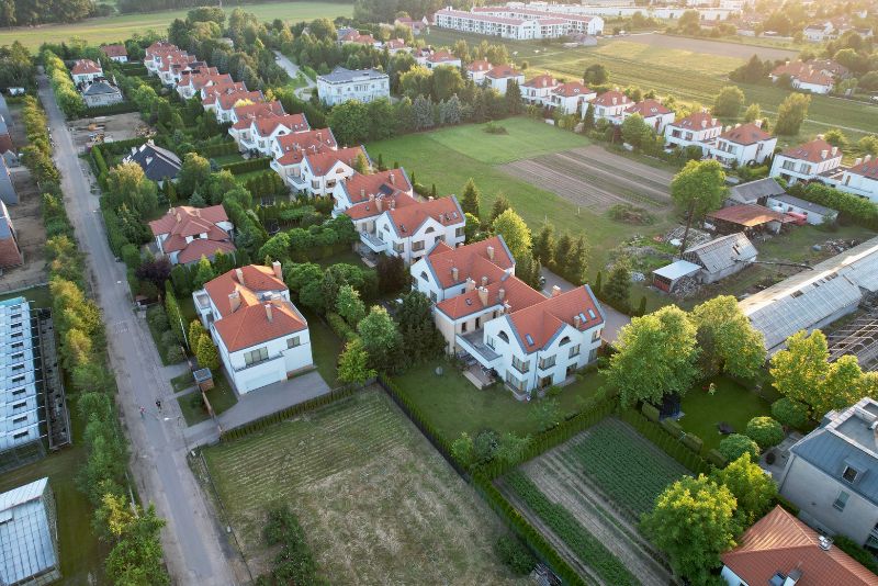 Widok z lotu ptaka na podmiejską okolicę z rzędami białych domów z czerwonymi dachami, zielonymi trawnikami, drzewami, ogrodami i pobliskimi polami uprawnymi.