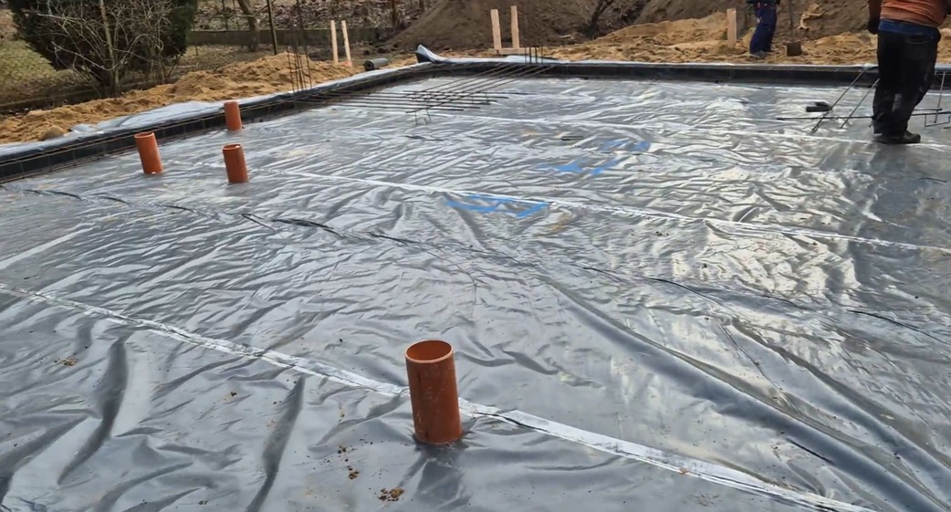 Fundamenty placu budowy przykryte dużą płytą plastikową, z kilkoma wystającymi pionowo pomarańczowymi rurami; w tle widoczna ziemia i narzędzia, wskazujące na trwające prace ociepleniowe płyty fundamentowej.