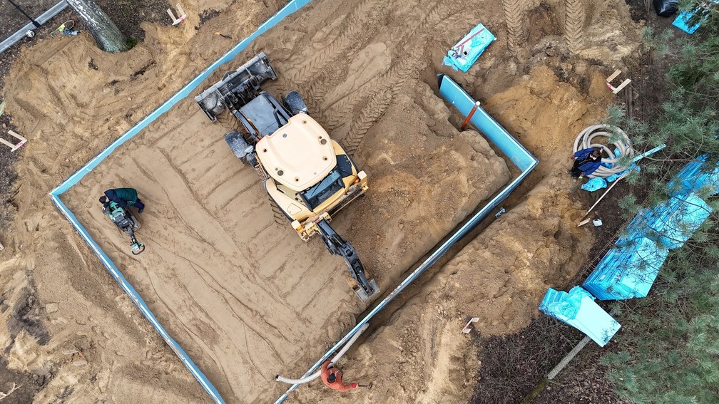 Widok z lotu ptaka na plac budowy, na którym widać spychacz przemieszczający piasek pod niebieskimi fundamentami; trzech pracowników nadzoruje proces ocieplania płyty fundamentowej, przy czym sprzęt i materiały są rozrzucone po całym terenie.