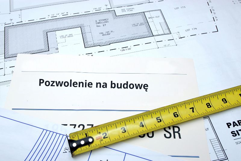 Polski dokument pozwolenia na budowę z napisem leży na planach architektonicznych darmowych projektów domów, z żółtą taśmą mierniczą umieszczoną ukośnie w poprzek dokumentów.